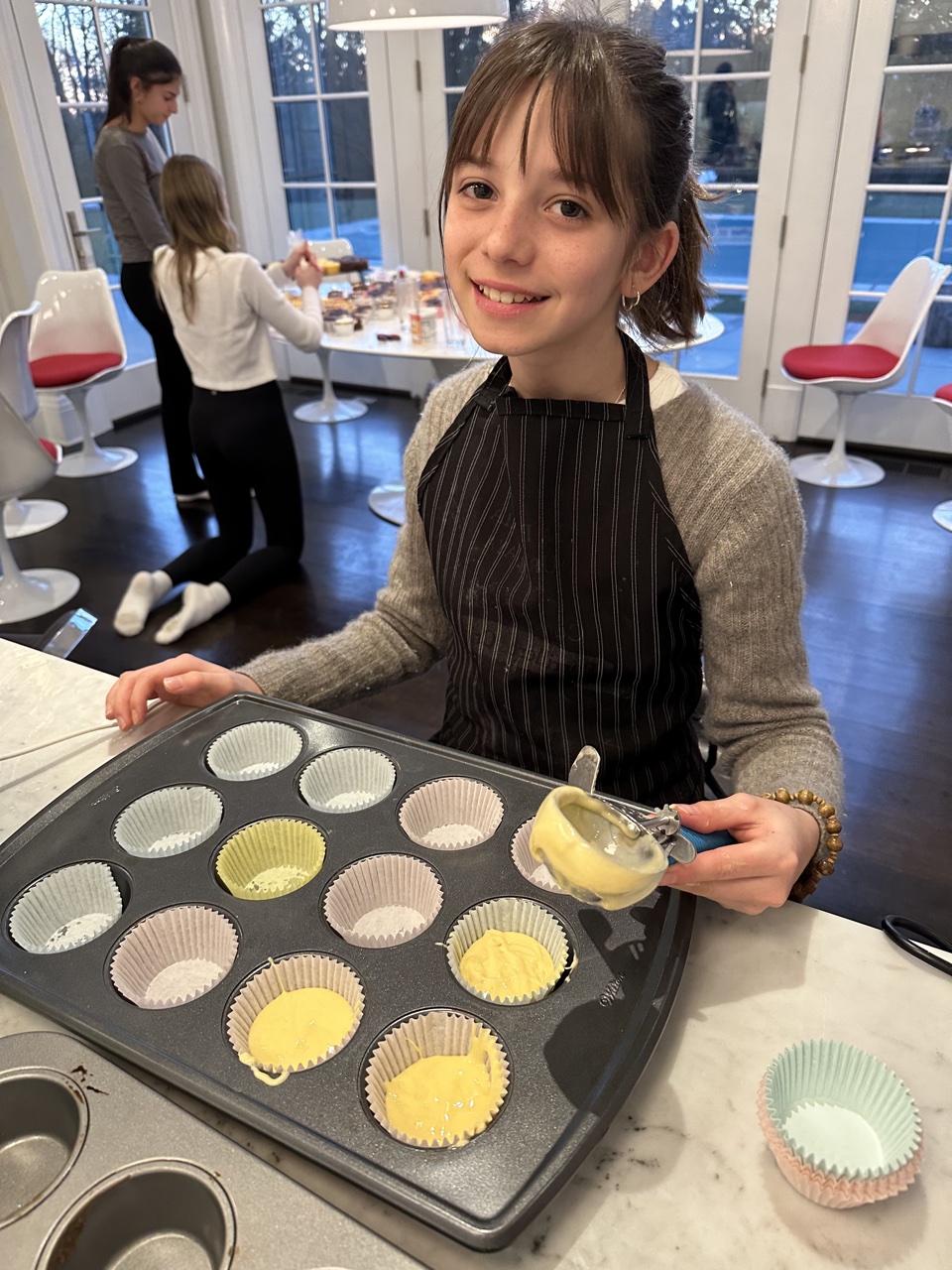 Young PeaceofCakes baker in a striped apron scooping cupcake batter into a muffin tin