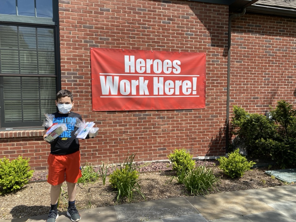 Young PeaceofCakes volunteer wearing a mask and holding gift-wrapped cakes beneath a Heroes Work Here banner