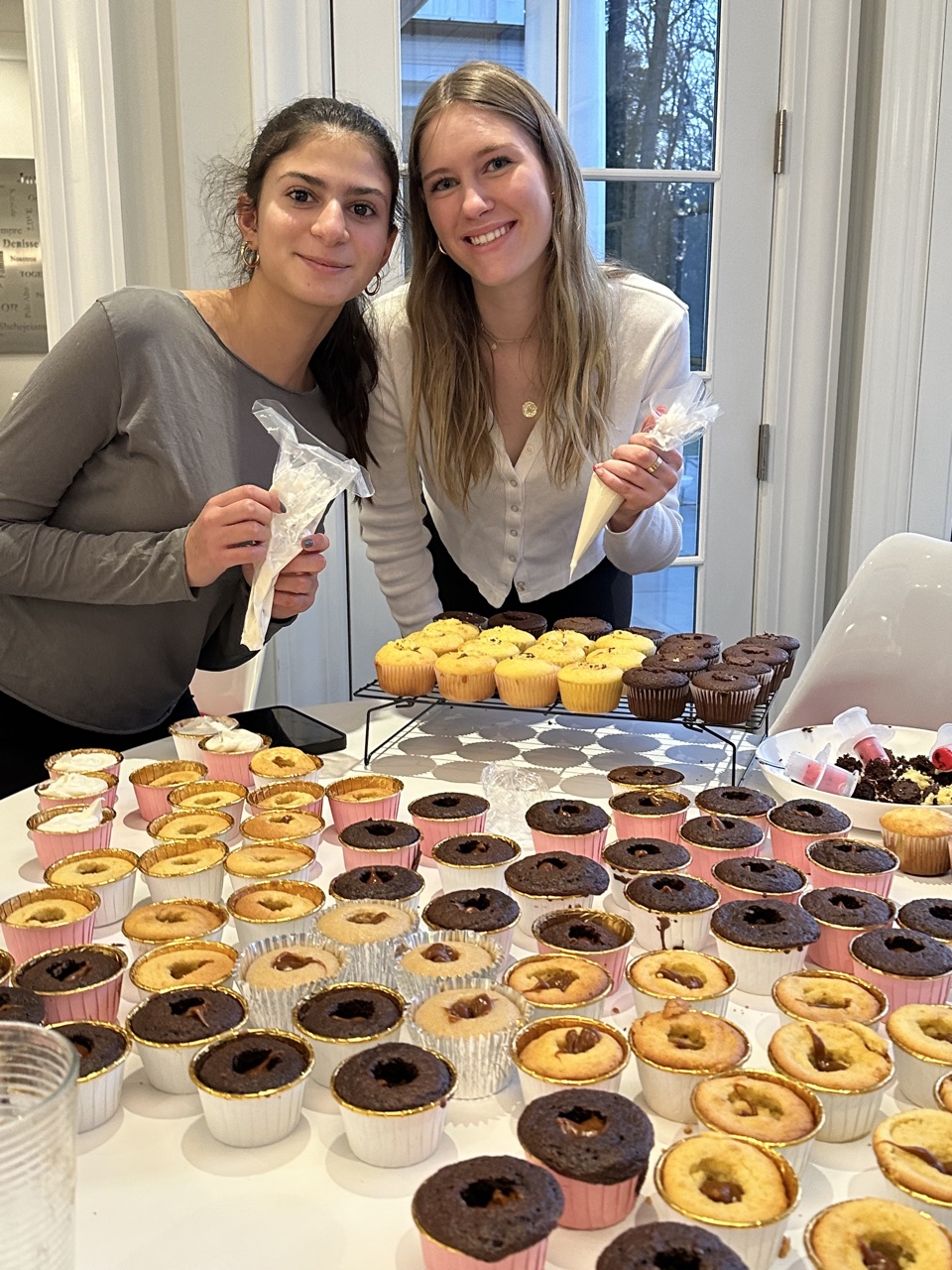 Two PeaceofCakes bakers holding piping bags, surrounded by cupcakes on a table
