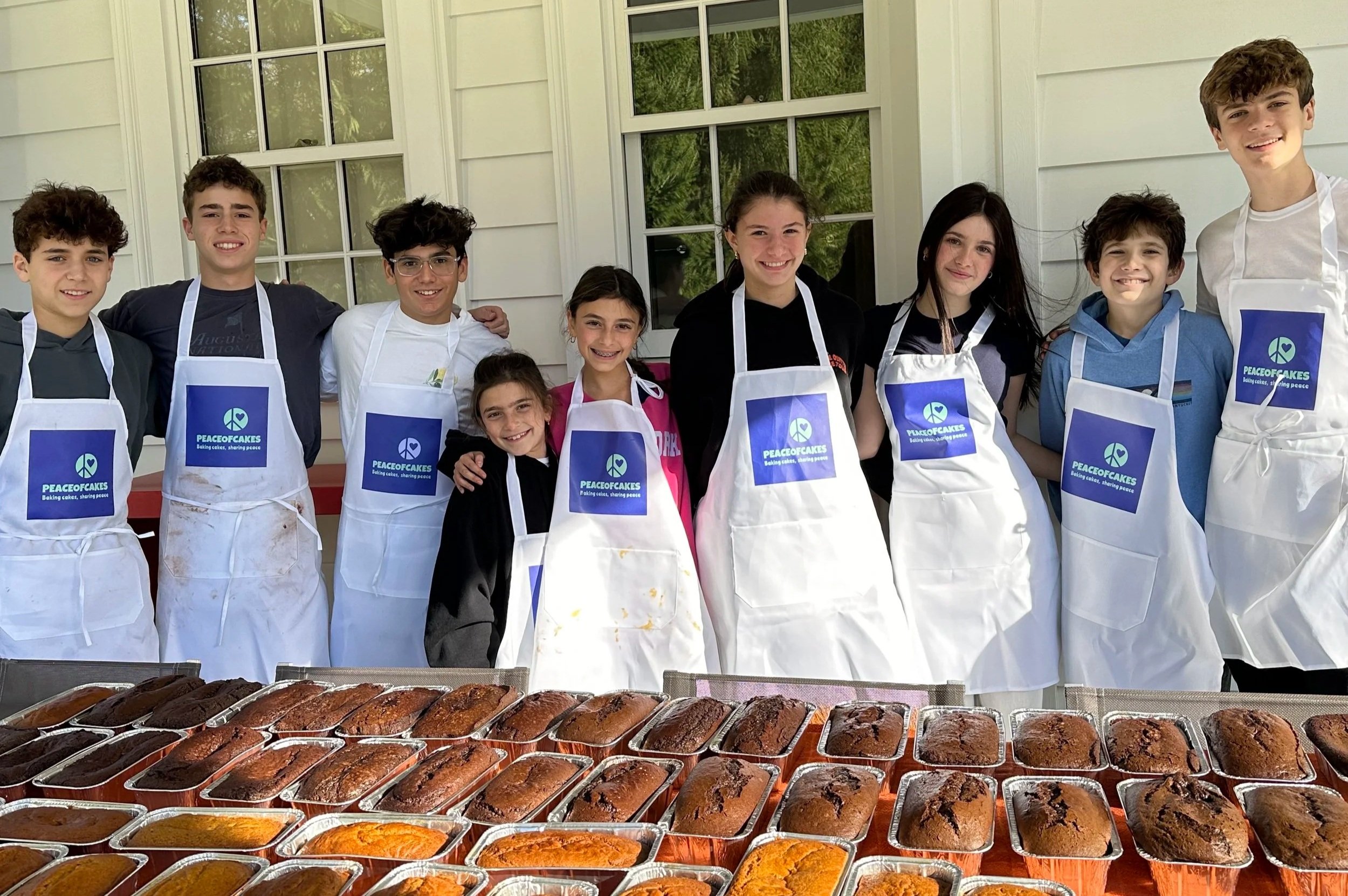 Nine young PeaceofCakes bakers standing behind rows of homemade cakes