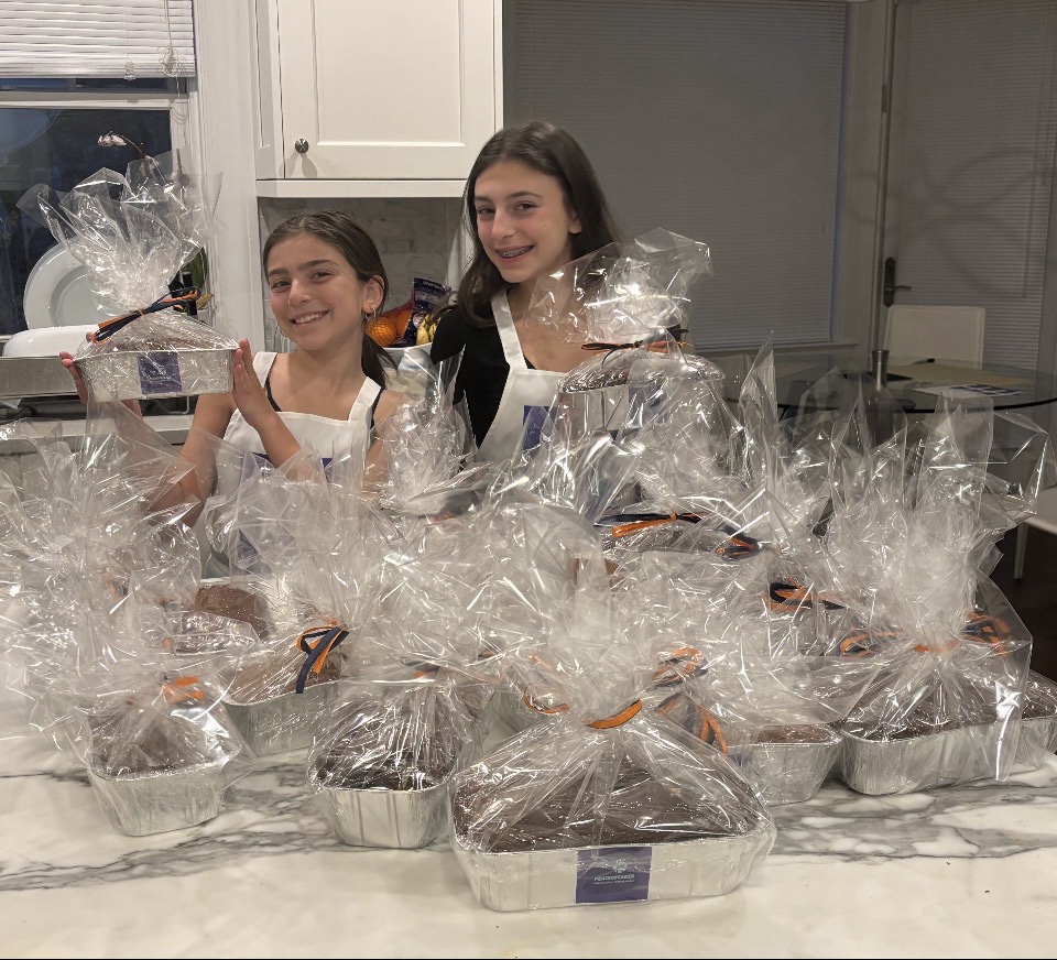 Two young PeaceofCakes bakers in aprons standing behind rows of gift-wrapped cakes